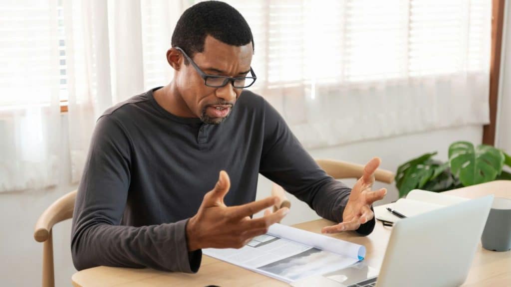 A Black man with glasses sits at a desk, gesturing and frowning at a document.