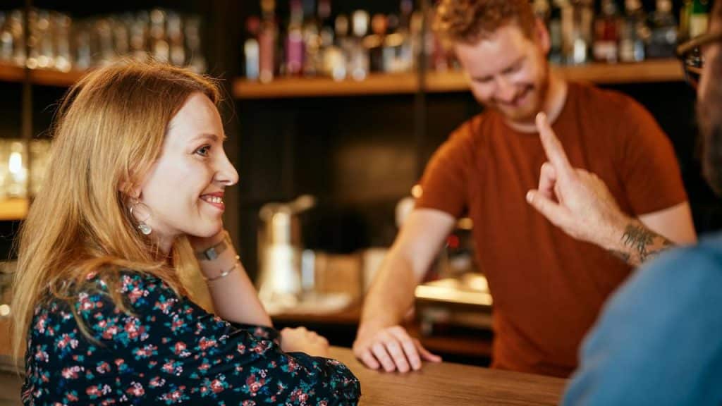 A smiling woman sitting at a bar counter while talking with someone next to her.