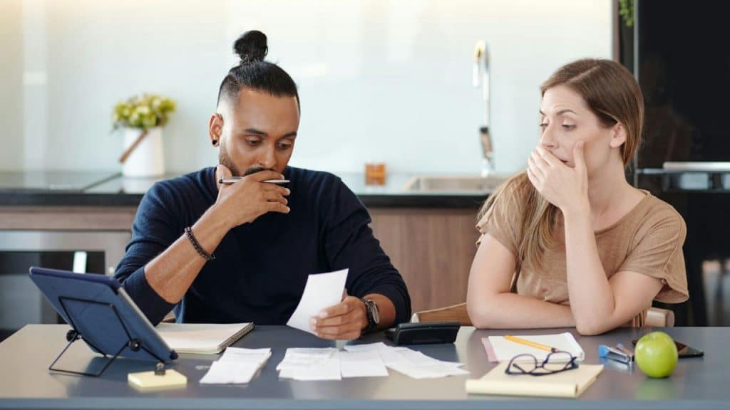 A confused couple looks at a pile of bills on their kitchen table.
