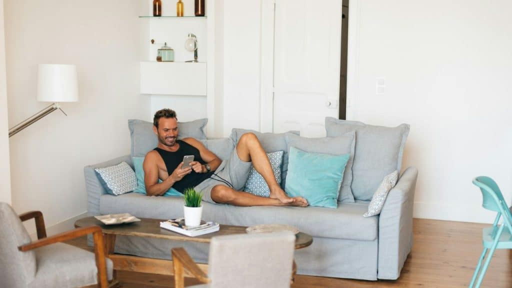 A man lies on a sofa in a living room, smiling at his phone.
