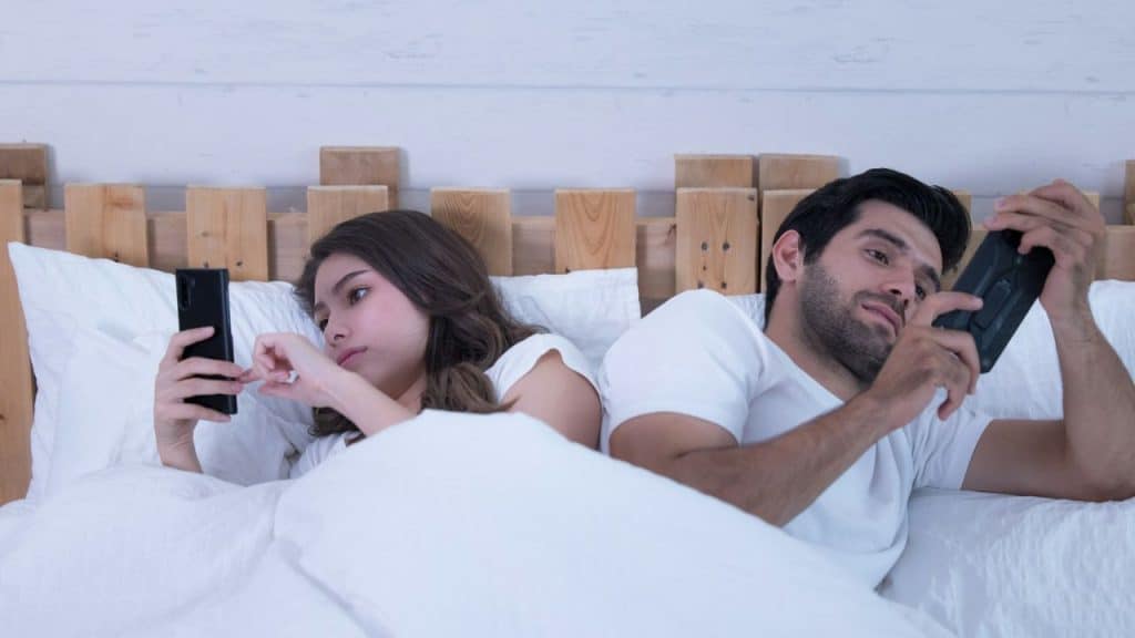 A man and woman lie in bed, back to back, looking at their phones.