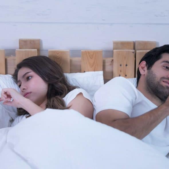 A man and woman lie in bed, back to back, looking at their phones.
