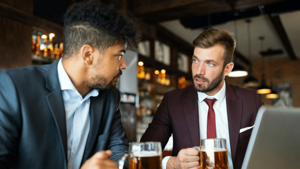 Two men in business suits sit at a bar with mugs of beer, looking at each other and talking.