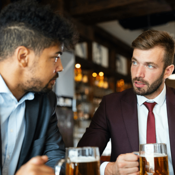 Two men in business suits sit at a bar with mugs of beer, looking at each other and talking.