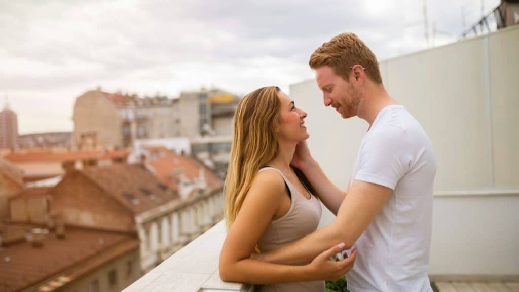 A smiling couple looks at each other while standing on a balcony with a city view.
