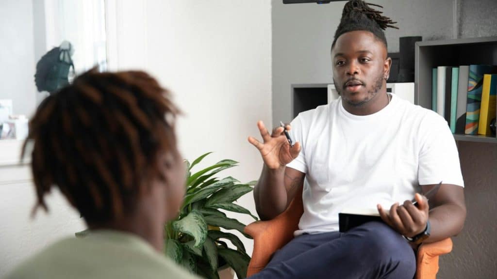 A man with dreadlocks sits in a chair, talking and gesturing to another person who is facing away from the camera.