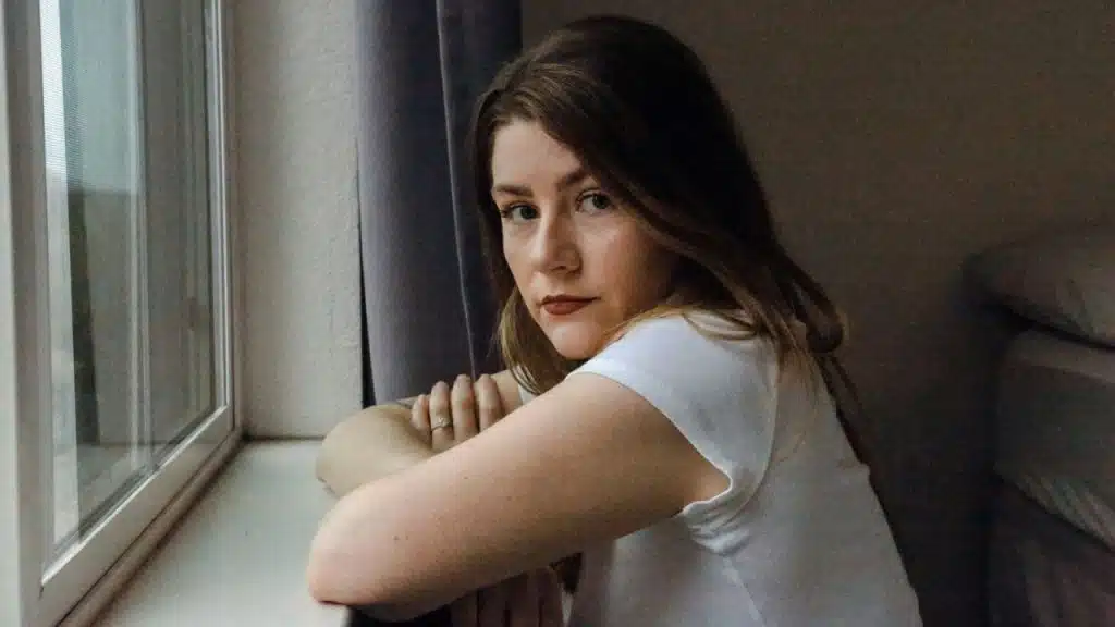 A woman with brown hair looks at the camera while leaning on a window sill.