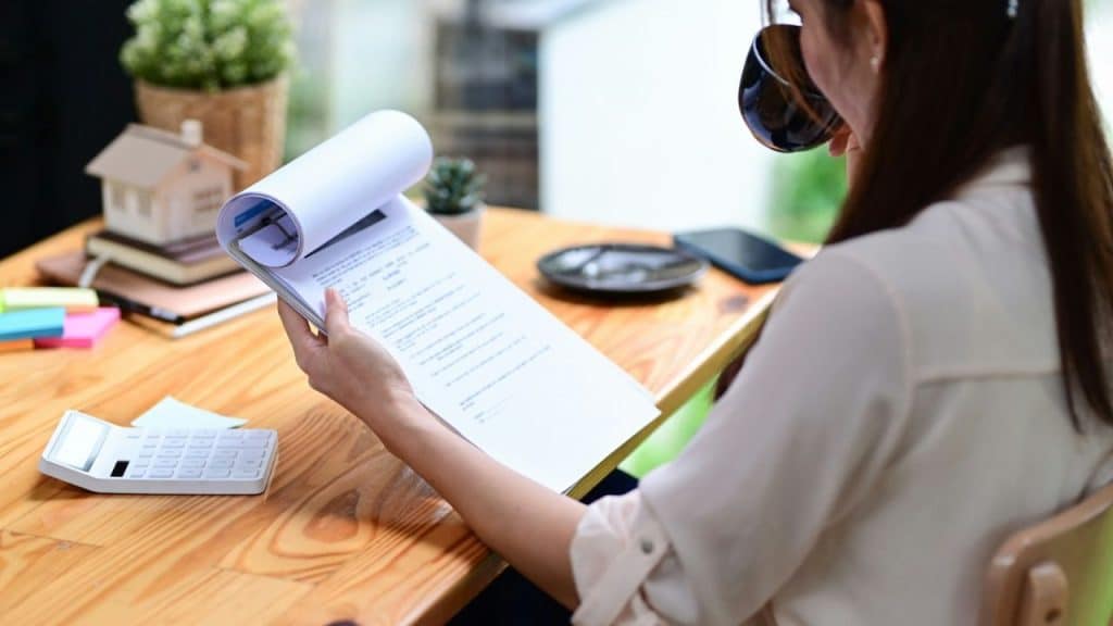 A woman is sitting at a desk and looking at a clipboard with papers on it.