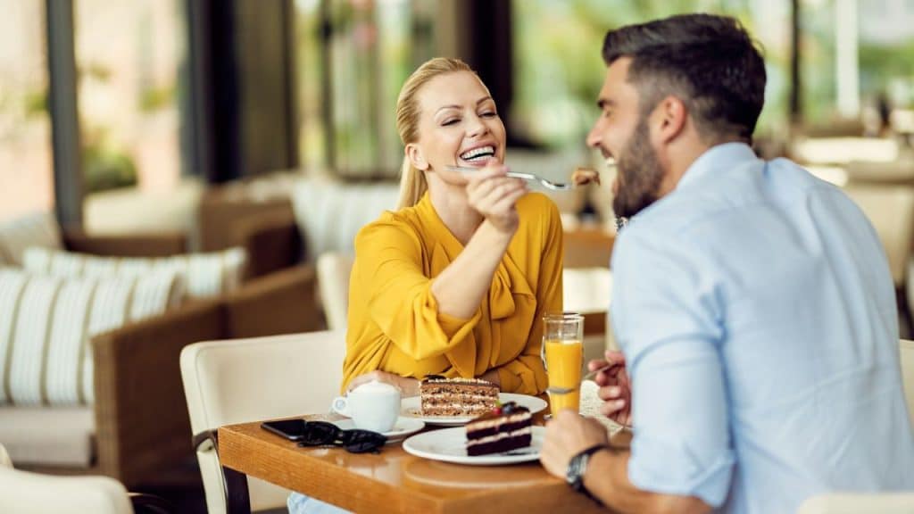 Blonde woman feeding dark-haired man cake with a fork at a restaurant table.