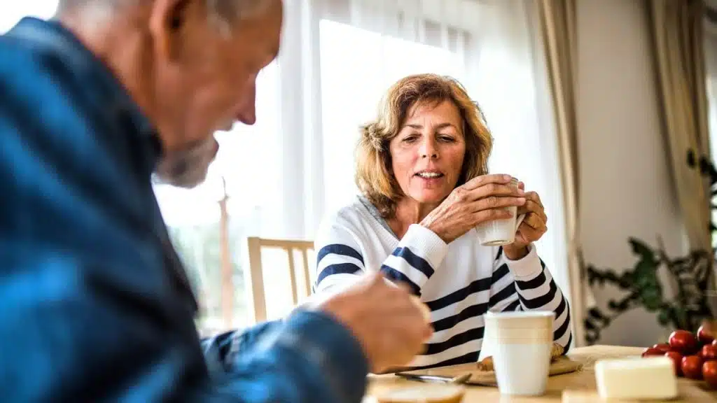 A smiling older couple sits at a dining table, drinking coffee and talking.
