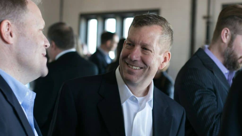 A middle-aged man in a dark suit and white shirt smiles while talking to another man in a crowded indoor setting.
