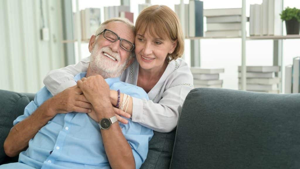 An elderly woman hugging a very happy, smiling man from behind on a couch.