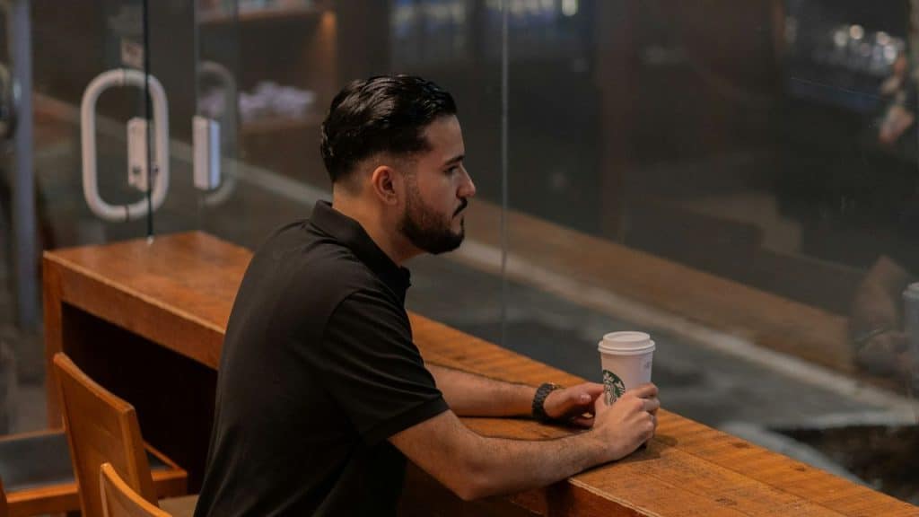 A pensive man with a beard sits alone at a coffee shop table.
