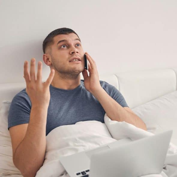 A man sitting in bed talking on the phone with a laptop in front of him.