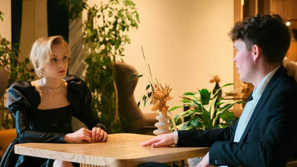A blonde woman and a man in a suit sit across a table in a cafe.
