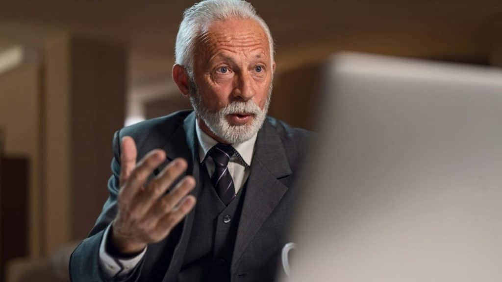 A gray-haired man in a suit gestures while looking at a computer screen.
