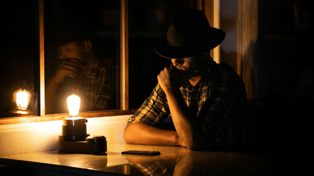 A pensive man in a plaid shirt and hat sits at a table, illuminated by a single lightbulb.