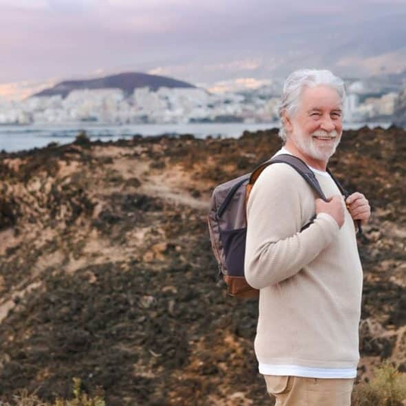 A cheerful elderly man with a backpack smiles at the camera on a rocky hillside overlooking a city and the sea.