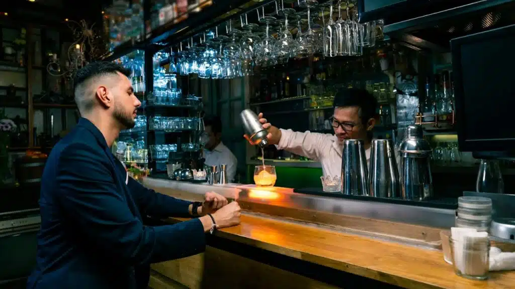 A man in a suit sits at a bar as a bartender pours a drink from a shaker.