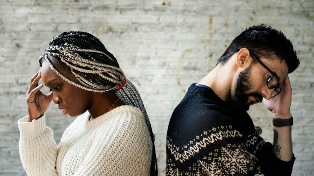 A man and a woman stand back to back, holding their heads in their hands.