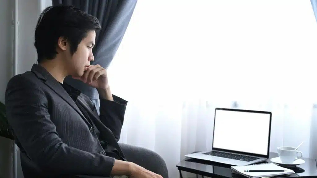 A serious man sits and stares thoughtfully at a laptop with a blank screen.