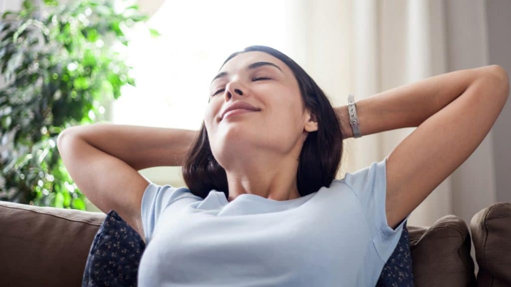A young woman is leaning back on a sofa with her eyes closed, smiling, and her hands behind her head.