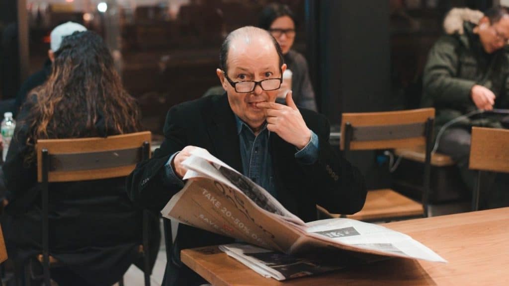 A surprised older man wearing glasses reads a newspaper at a cafe.