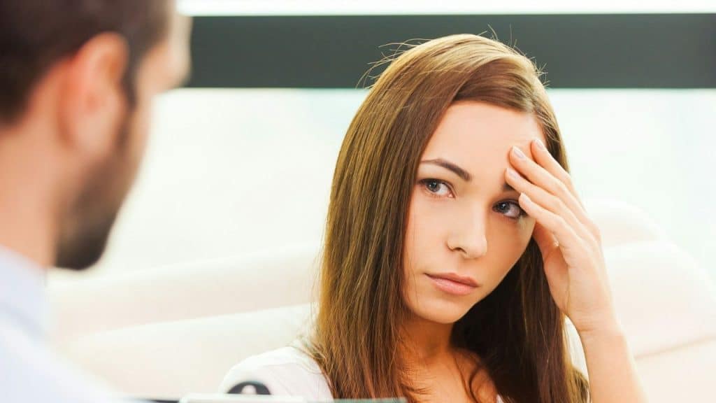 A woman with a worried expression is sitting across from a man, with her hand on her forehead.