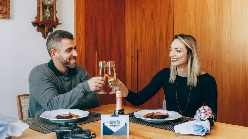 A couple smiles and clinks champagne glasses over a romantic dinner at a wooden table.