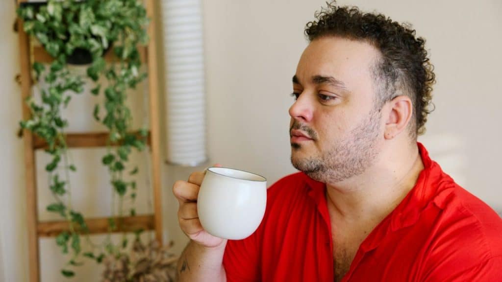 A man in a red shirt holds a white mug and looks off to the side indoors.