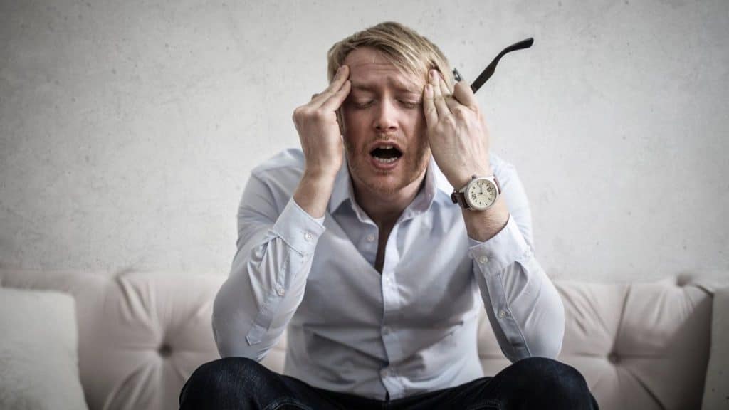 A blond man sitting on a couch, holding his head with both hands, looking stressed.