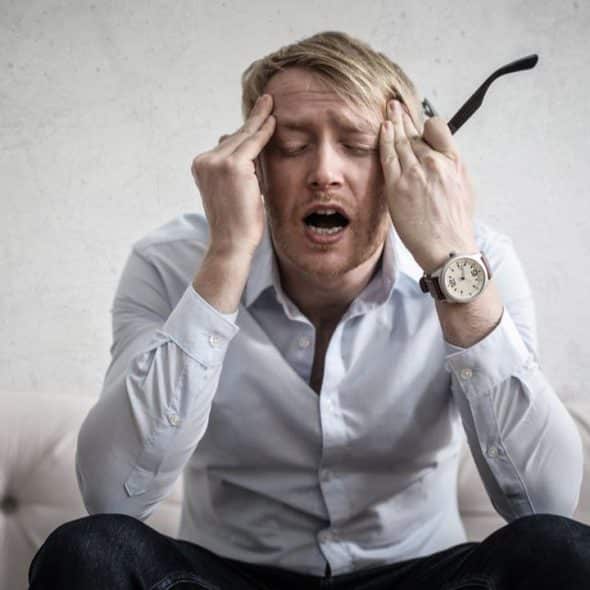 A blond man sitting on a couch, holding his head with both hands, looking stressed.