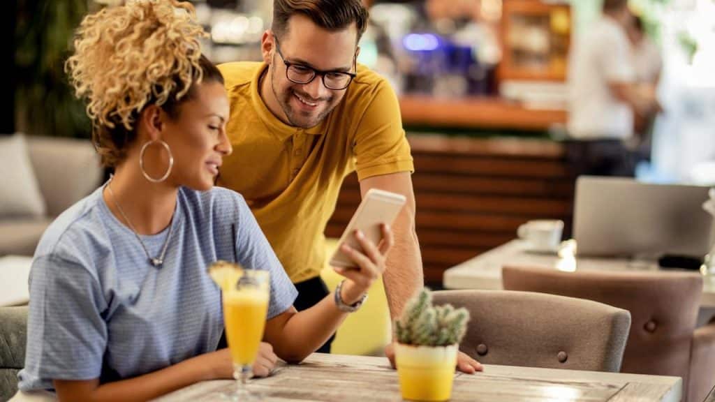 A woman showing her phone to a smiling man at a café table with drinks.