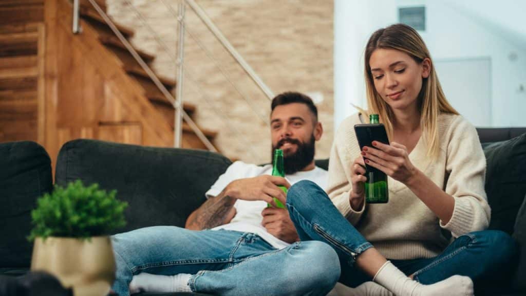 A distracted woman looks at her phone while a man sits beside her, holding a beer.