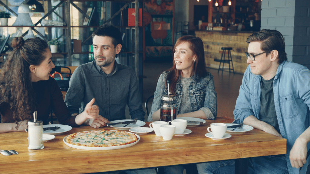 A man in glasses looks on as a couple at a table talks animatedly with their friends.