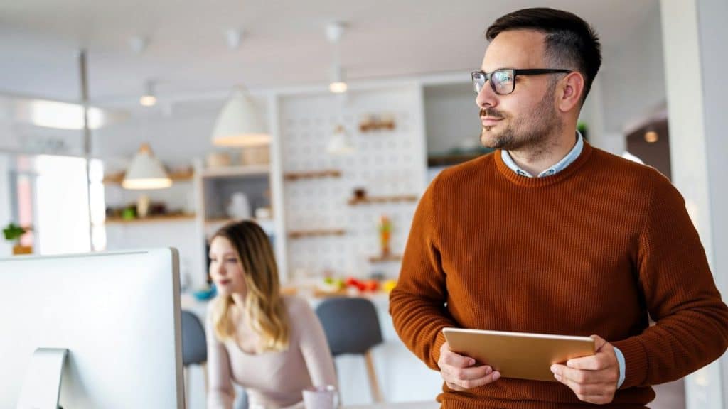A man holding a tablet looks to the side while a woman works at a computer in the background.