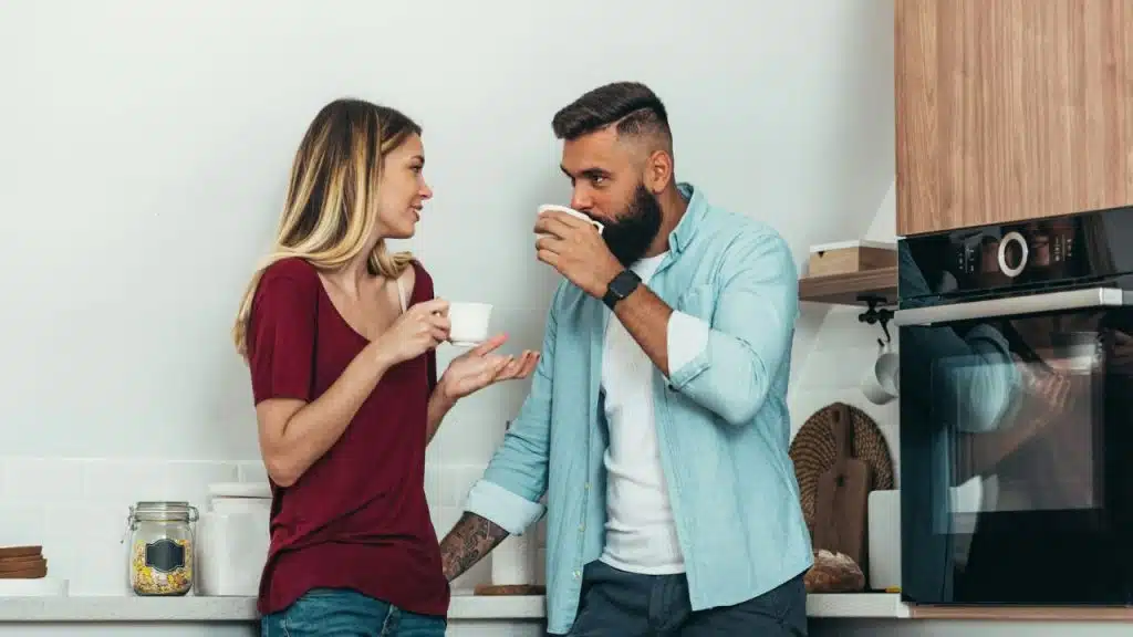 A couple is in a kitchen, drinking coffee. The man with a beard is taking a sip from his mug, while the woman is gesturing with her hands as if she is talking.