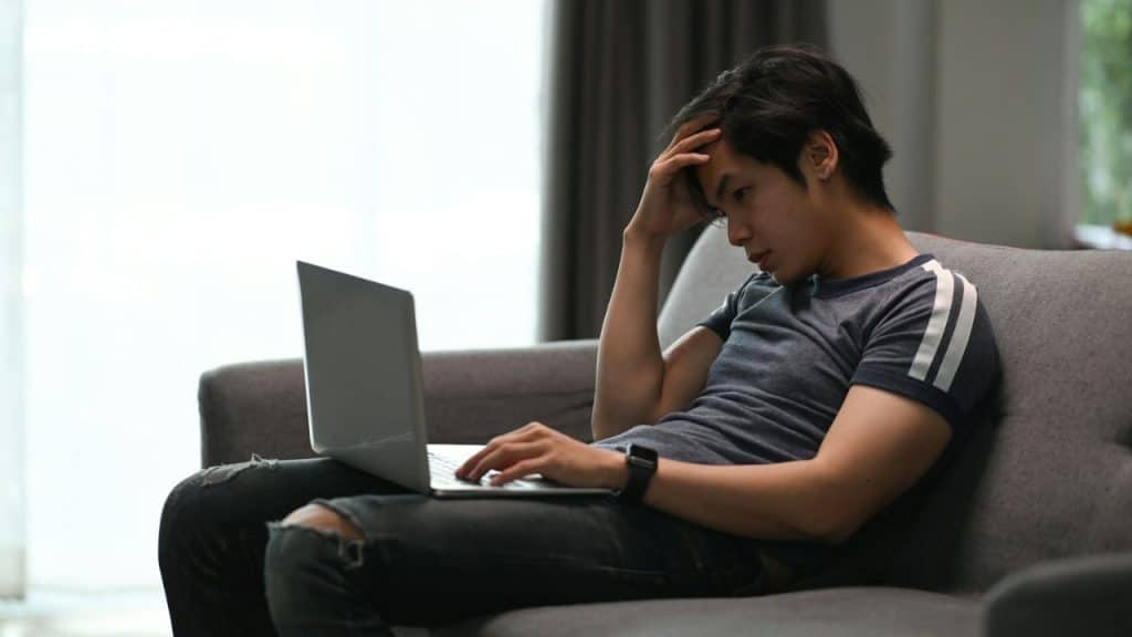 A man sits on a sofa, looking at his laptop and holding his head in his hand.