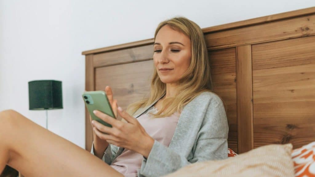 A woman with blonde hair sits in bed, looking at her phone.