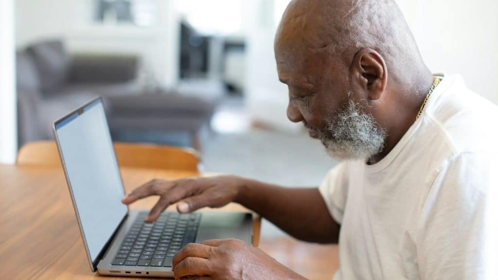 A gray-bearded older man wearing glasses uses a tablet outdoors.