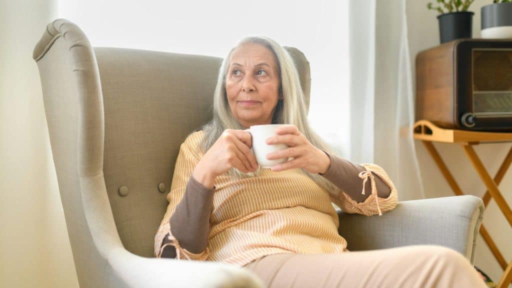 An older woman with long gray hair is sitting in an armchair, holding a mug and looking to the side.
