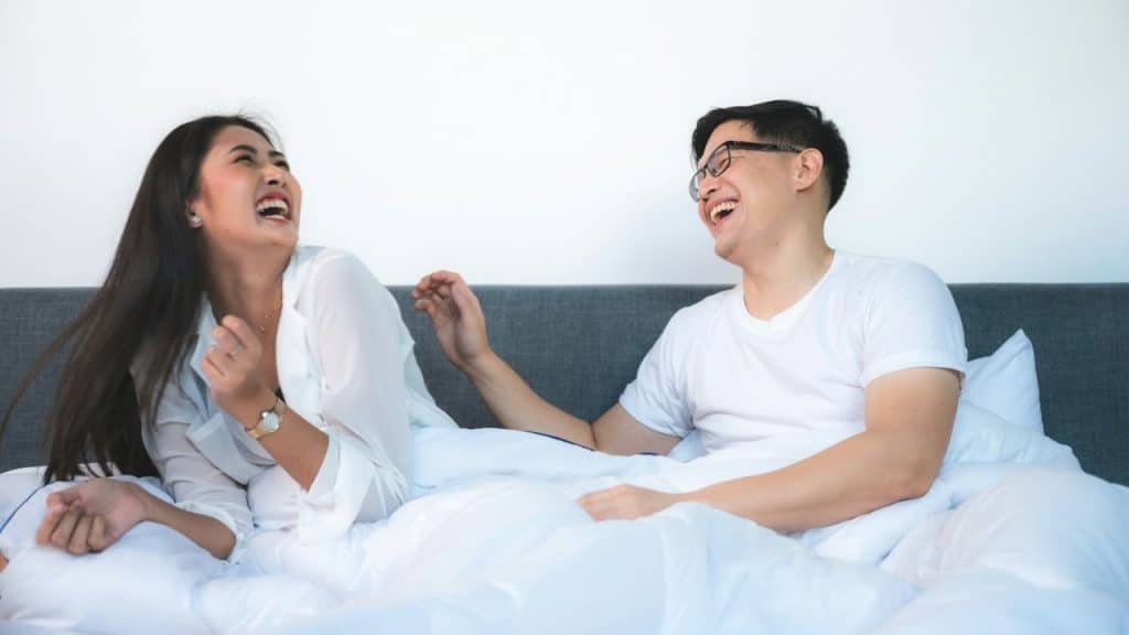 Asian man and woman laughing loudly while sitting up in a bed with white bedding.