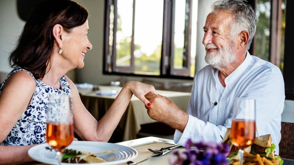 A happy, smiling, older couple is at a restaurant, holding hands across the table.