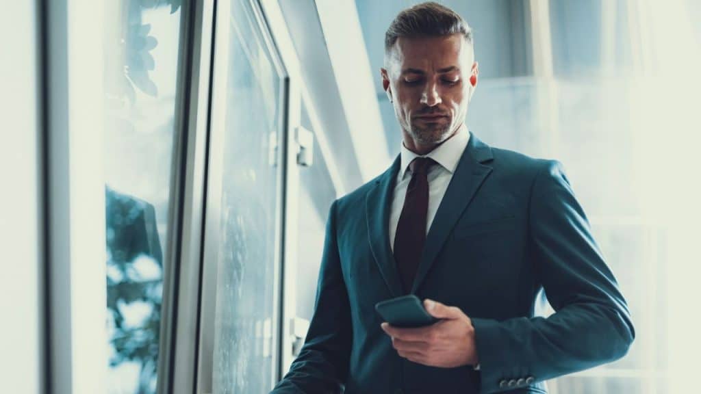 A businessman in a blue suit and tie looks at his phone while walking indoors.