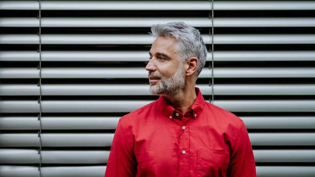 A confident, silver-haired man in a red shirt stands against a striped background.