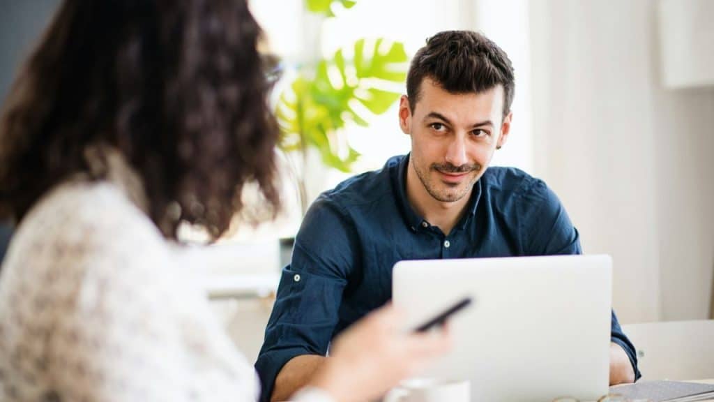 A smiling man looks past his laptop toward a person with dark, curly hair in the foreground.