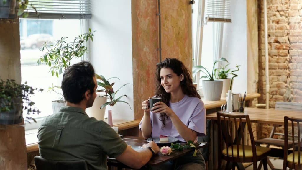 A woman holding a mug and smiling while talking with a man at a café table.