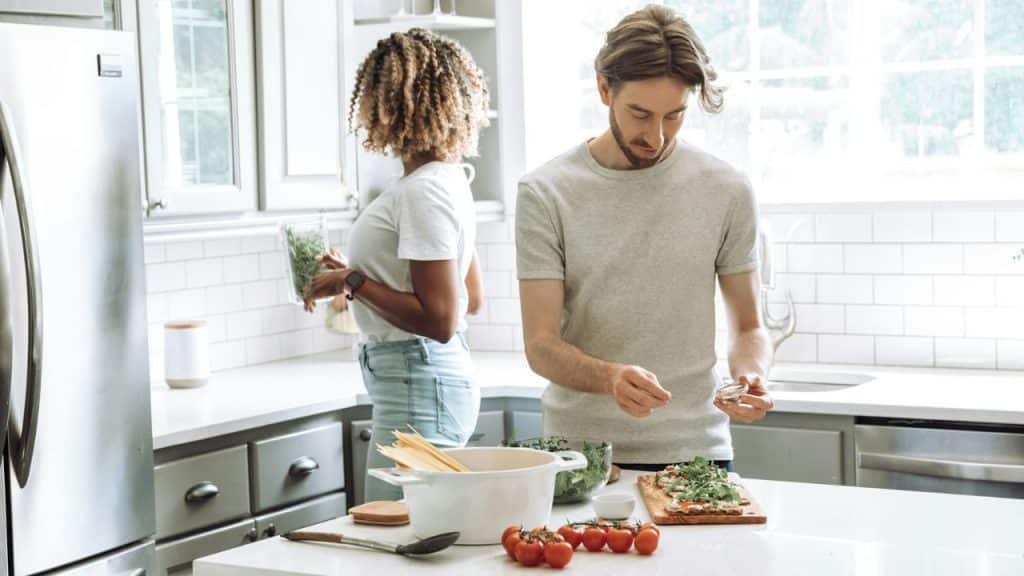 A multiracial couple in a kitchen, one preparing food while the other stands nearby.