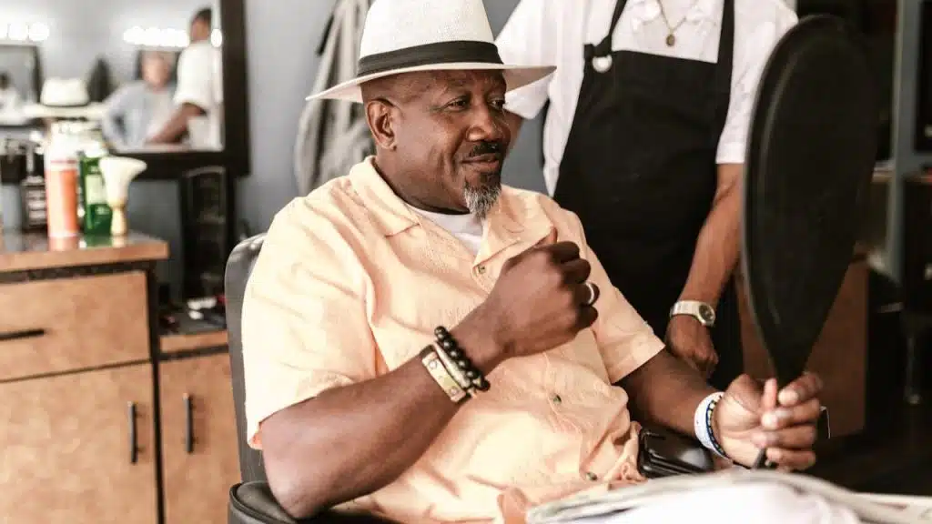 A smiling man with a goatee and a white fedora sits in a barber's chair, holding up a mirror.