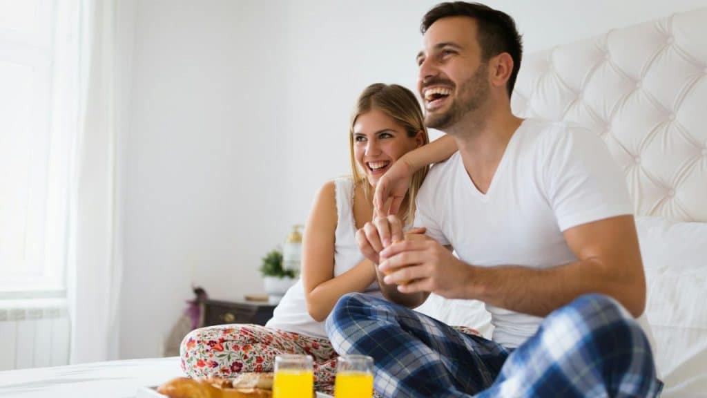 A happy couple in pajamas sits on a bed, laughing and holding hands. A breakfast tray is in front of them.